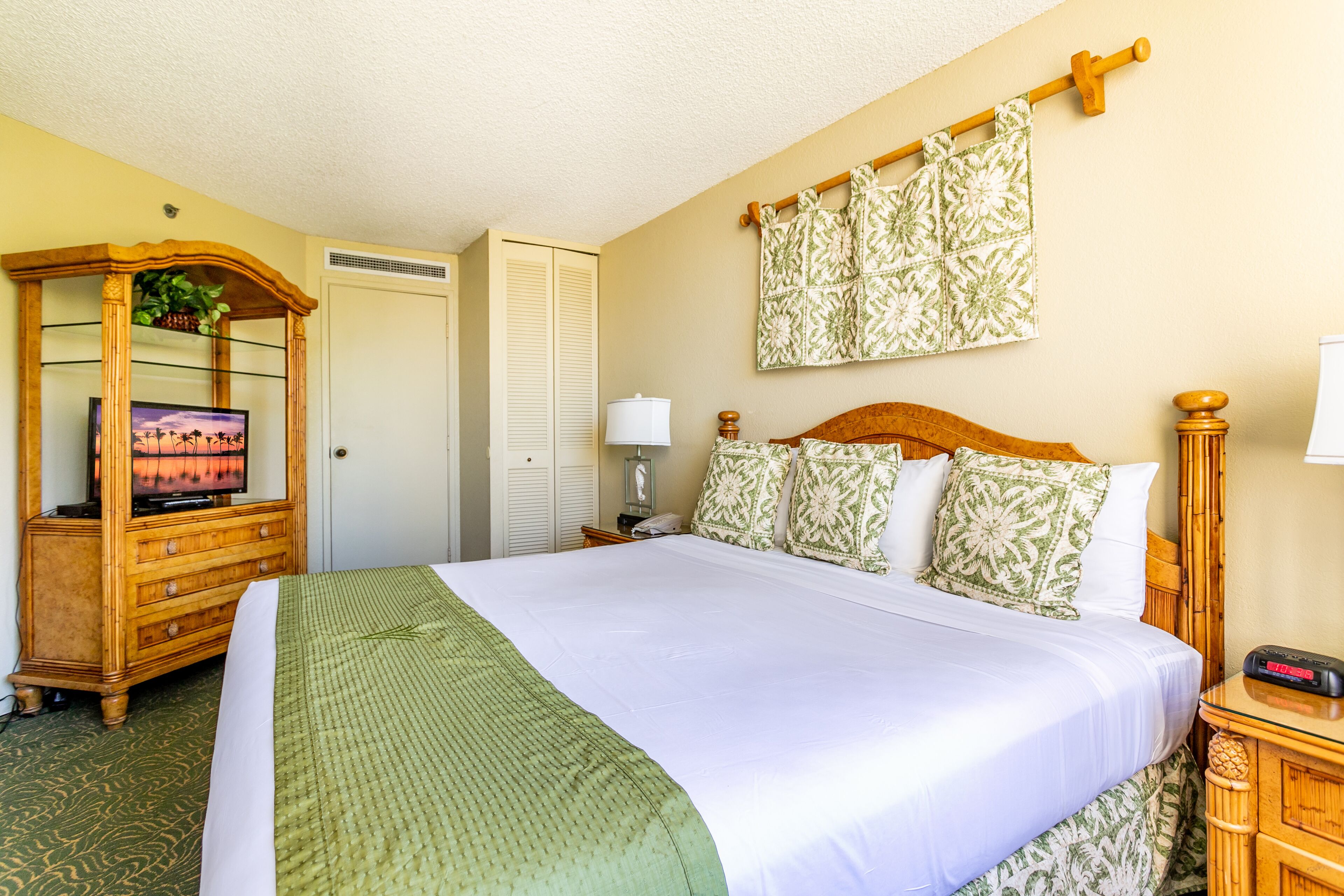 A hotel room with a large bed, wooden headboard, a wooden dresser with a TV, a bedside table with a lamp, and a wall-mounted towel rack.