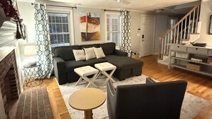 Ett vardagsrum med en soffa, en pall och en fåtölj, en öppen spis och en trappa.
