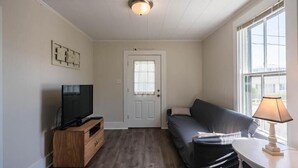 A small, well-lit room with a wooden entertainment unit, a black sofa, a small table with a lamp, and a large window providing natural light. - Salty Air Suites, B:  Easy to walk in, One level, 3 beds, ocean views. (Old Orchard Beach)