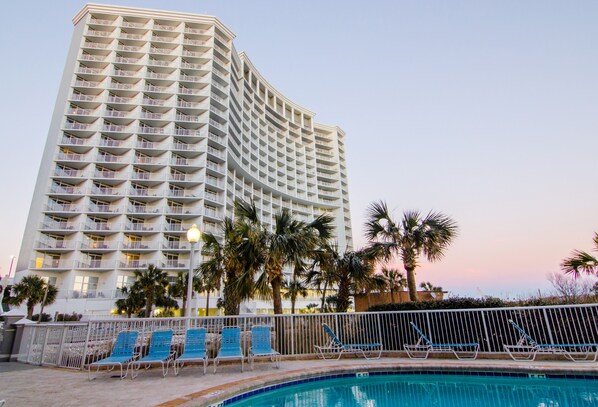 A high-rise hotel with multiple balconies, a swimming pool with lounge chairs, and palm trees.
