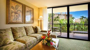 Ein Hotelzimmer mit einer Couch, einem Couchtisch mit Blumenarrangements, einer Lampe und einem Balkon mit Blick auf Palmen und Gebäude.