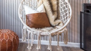 A macramé hanging chair with a fur throw, a brown leather pouf, and a cactus painting on the wall.