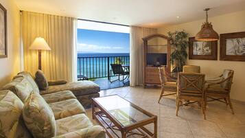 A living room with a sofa, coffee table, and wicker chairs, a television, and a large window with a view of the ocean.