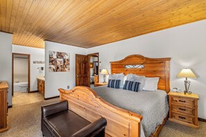 Une chambre à coucher avec un lit en bois, une table de chevet, une chaise et une salle de bain visible à travers une porte ouverte.