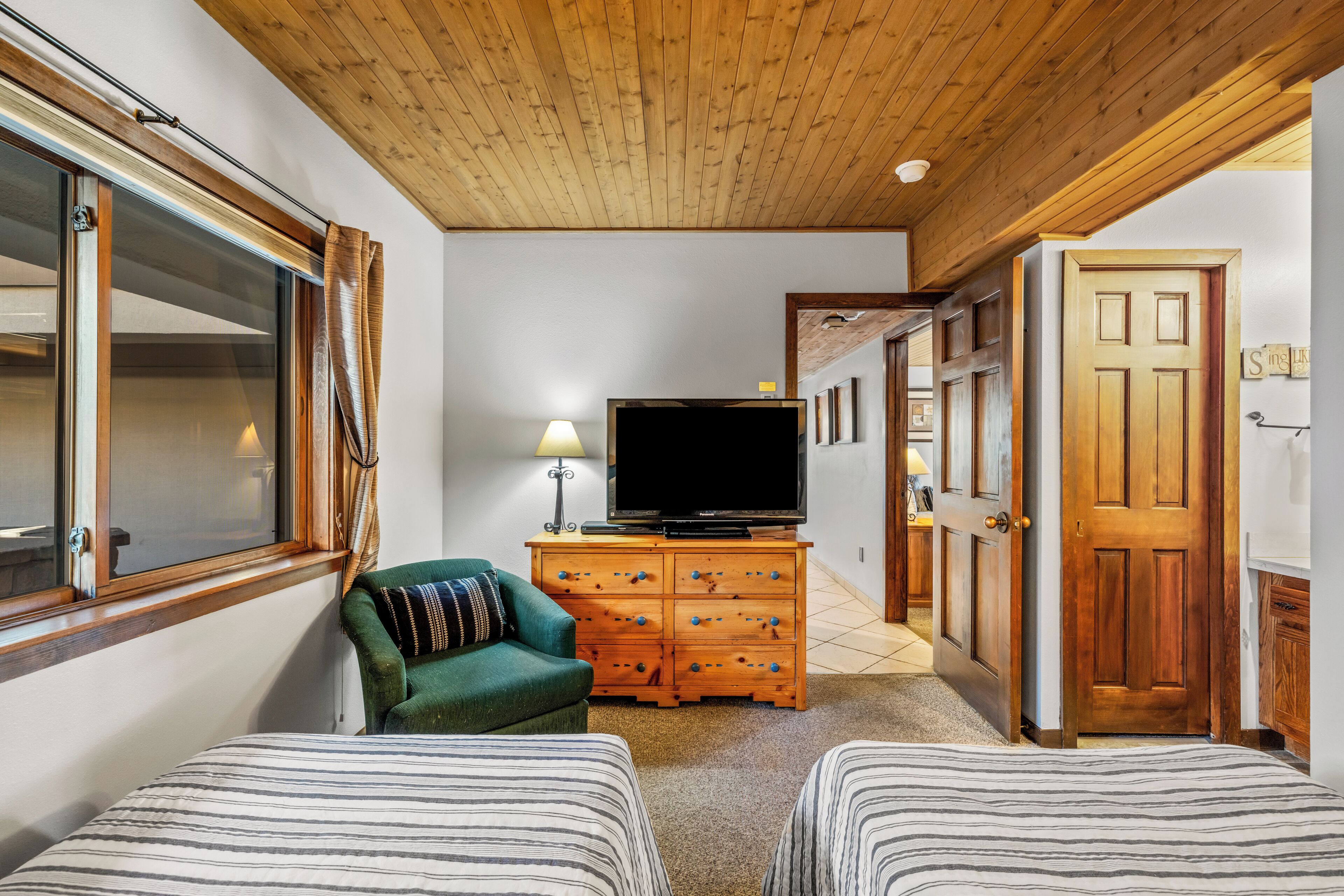 A cozy room with a wooden ceiling, a television on a wooden dresser, two striped beds, and a green armchair.