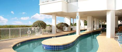 A swimming pool with a curved edge, a white building with balconies, and a clear blue sky.