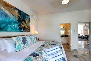 A hotel room with a large abstract painting, a bed with floral bedding, a neatly made bed with towels, a kitchen area visible through an open door, and a ceiling fan.