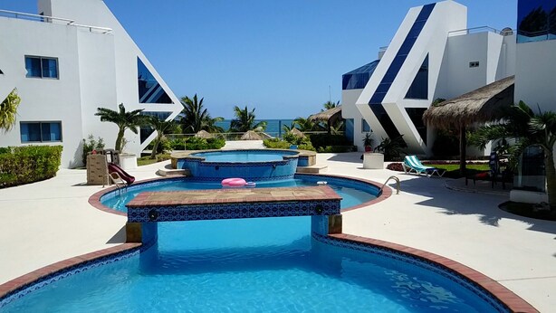 A pool area with multiple pools, palm trees, and a view of the ocean. - Oceanfront Luxury Townhome Condo Wifi 3 Smart TV'S Pool & Spa (Puerto Morelos)