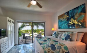 A bedroom with a bed, a television, a ceiling fan, a painting, and a view of the outdoors.
