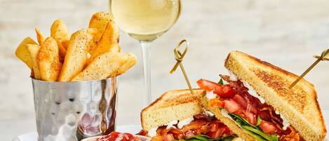 A sandwich with bacon, tomato, and lettuce, a glass of white wine, and a container of fries.