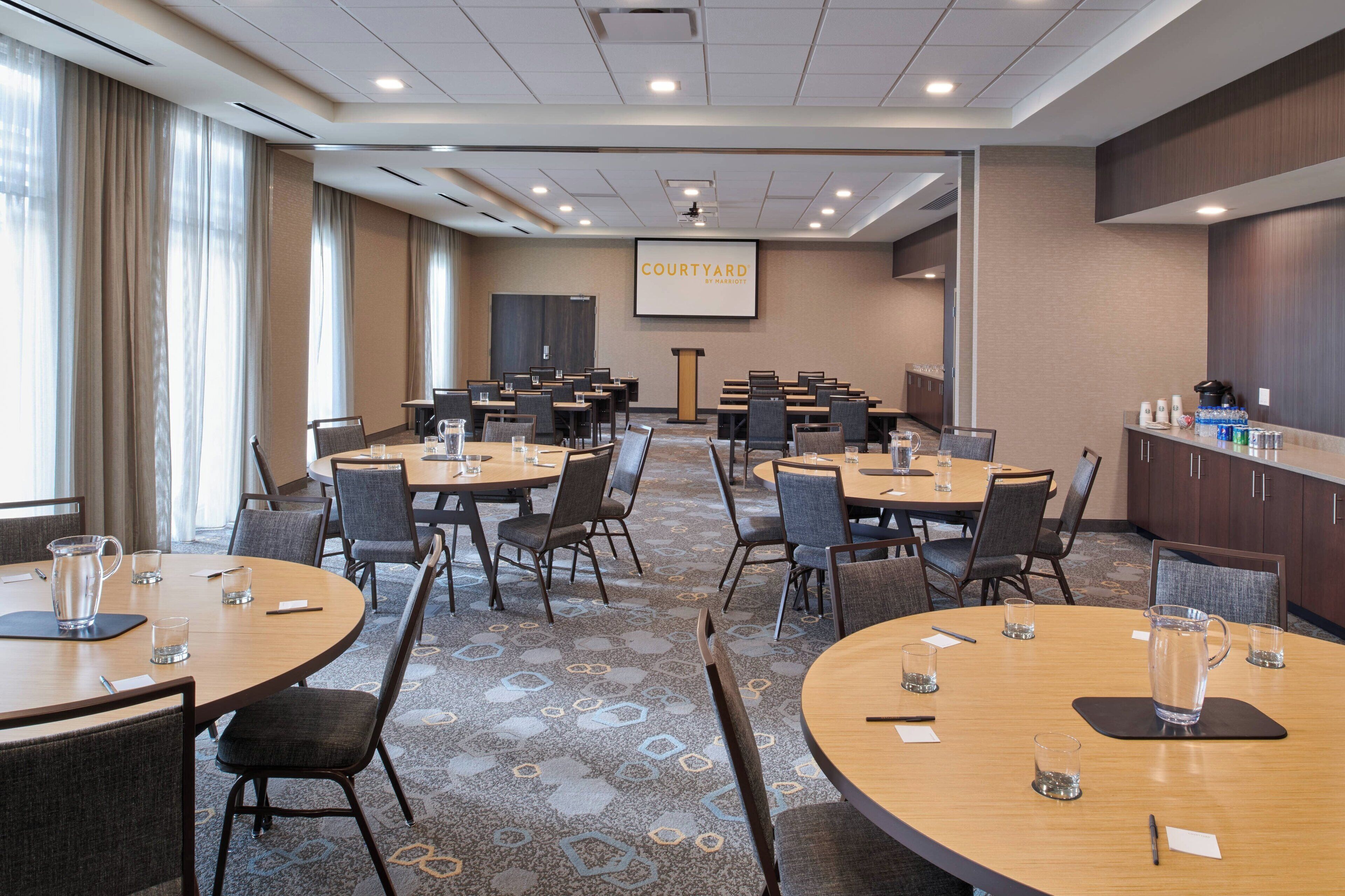 Une salle de conférence équipée de plusieurs tables et chaises, d’un grand écran affichant le logo « COURTYARD BY MARRIOT », et d’un coin cuisine.