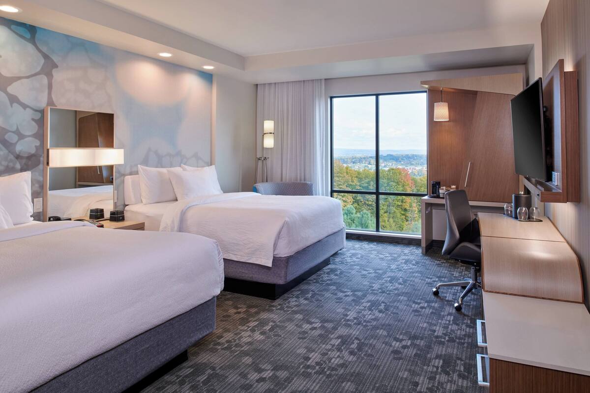 A hotel room with two beds, a desk, a chair, a TV, and a large window offering a city view.