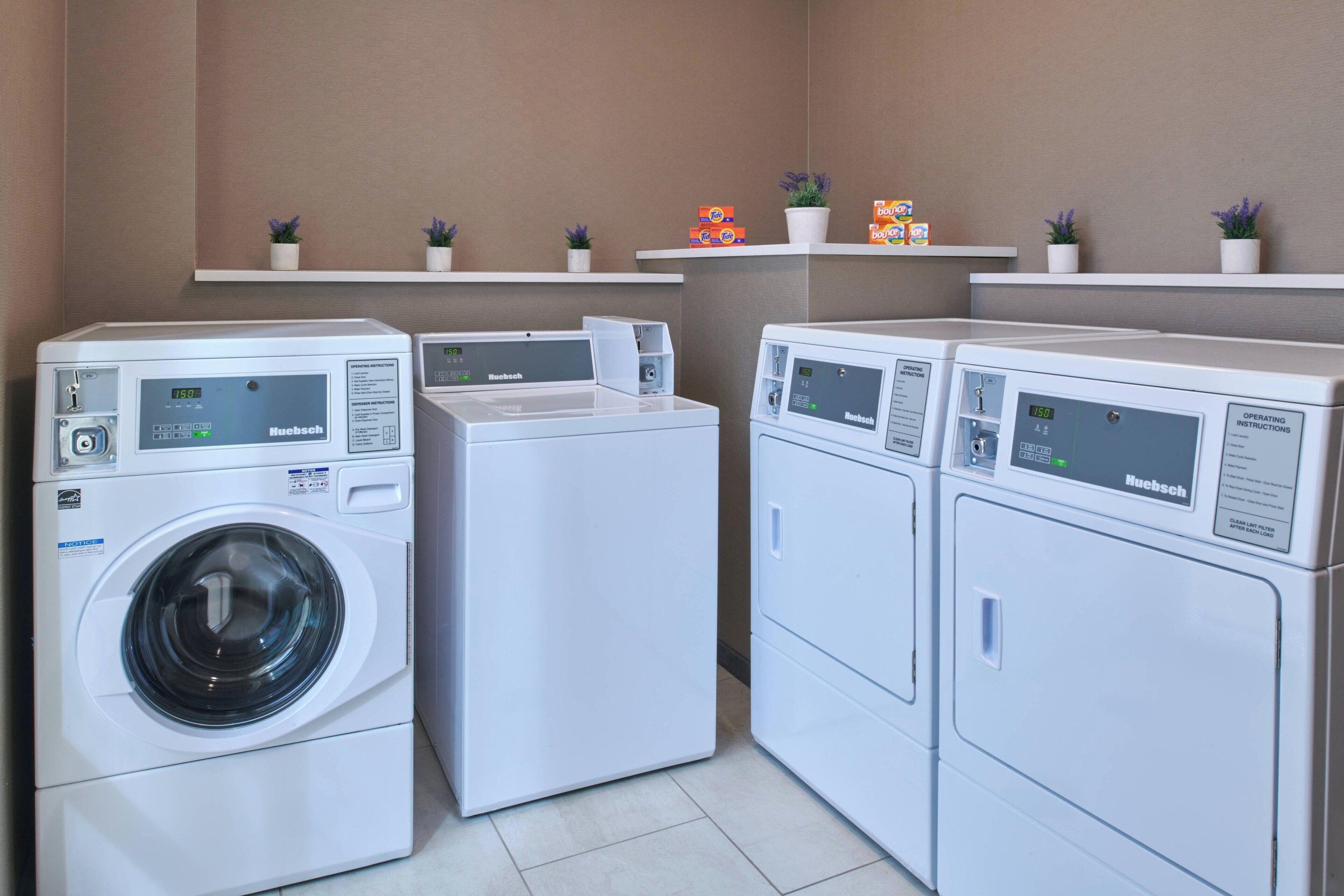 A laundry area with four white washing machines, each with a control panel. There are small potted plants on shelves above the machines.