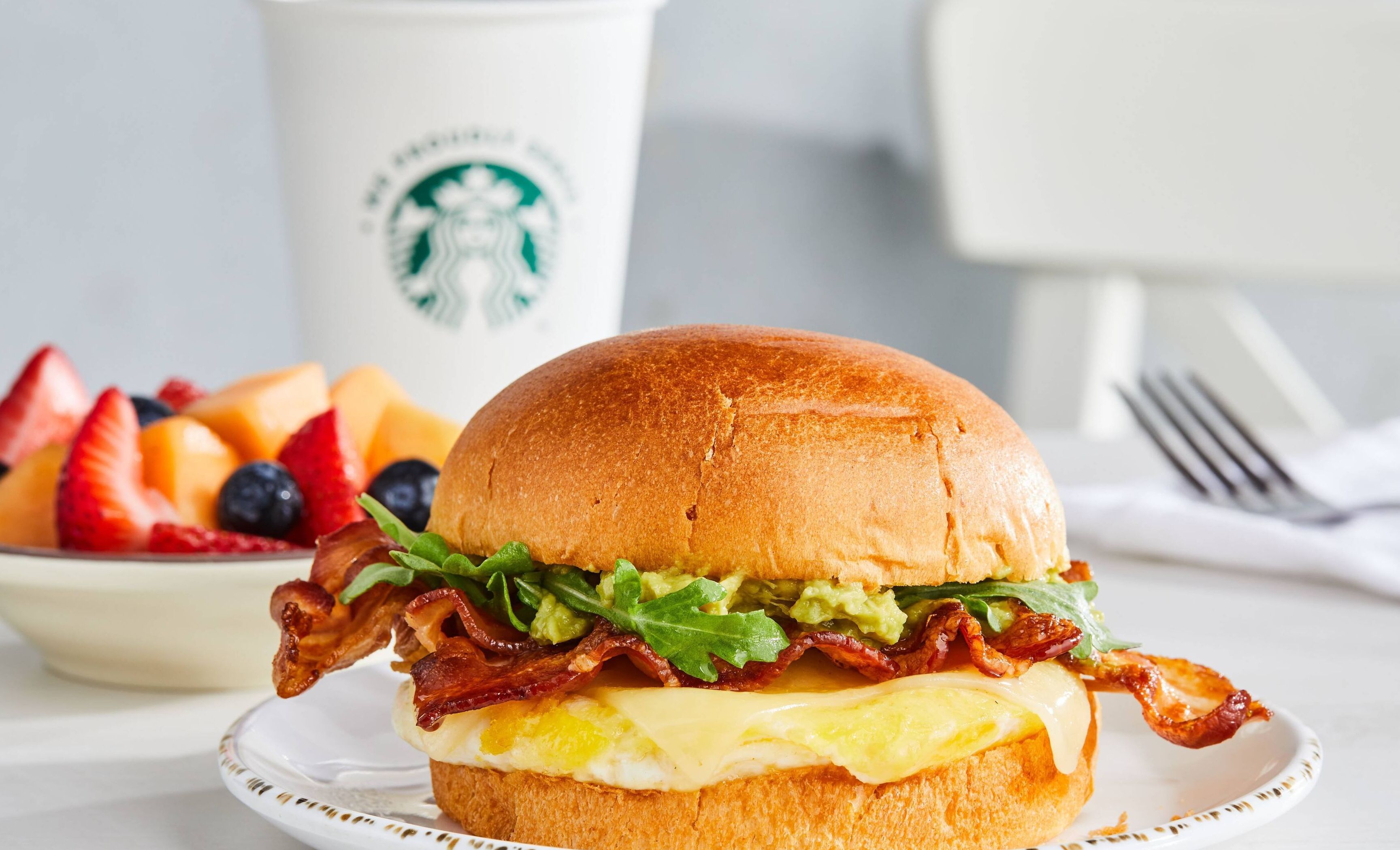 Un sándwich de desayuno con tocino, lechuga y huevo en un pan, un bol de fruta y una taza de Starbucks al fondo.