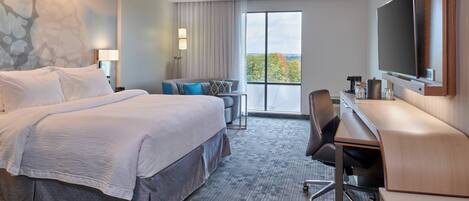 A hotel room with a large bed, a desk, a chair, a TV, and a view of the outdoors.