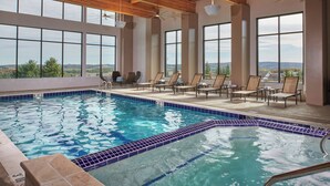 An indoor swimming pool with clear blue water, surrounded by lounge chairs and large windows offering a view of the landscape. - Courtyard by Marriott Petoskey at Victories Square (Petoskey)