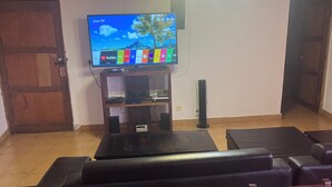 Een woonkamer met een televisie op een houten standaard, een zwarte poef en een rode fauteuil.