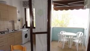 Une cuisine compacte équipée d’un lave-linge, un coin repas avec une table et des chaises, et un balcon avec vue sur les arbres.
