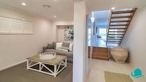 A modern living room with a sofa, coffee table, and artwork. A staircase leads to an upper level with a view. - The Sanctuary By The Sea – family and Pet Friendly, walk to the Beach (Catherine Hill Bay)