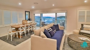 A spacious living area with a sofa, dining table, and chairs. There is a balcony with a view of the landscape. - The Sanctuary By The Sea – family and Pet Friendly, walk to the Beach (Catherine Hill Bay)