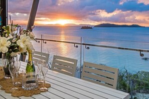 A table set for two with a bottle and glasses, overlooking a sunset over the sea. - Hidden Cove 6, Stunning Seaview Apartment on Hamilton Island (Hamilton Island)