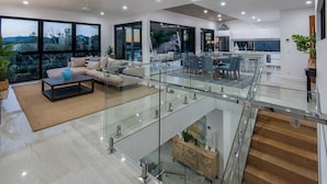 A modern, open-plan living area with a glass staircase connecting two floors, a dining area, and a kitchen. - Hidden Cove 6, Stunning Seaview Apartment on Hamilton Island (Hamilton Island)