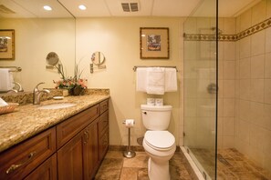 Baño con lavamanos de encimera de granito, armarios de madera, inodoro y ducha con mampara de vidrio.