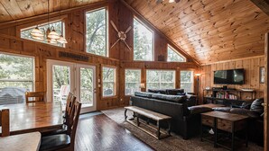A spacious living area with a wooden ceiling, large windows, a sofa, a television, and a dining table with chairs.
