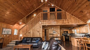 A spacious living area with a high ceiling, wooden walls, a staircase leading to an upper level, a kitchen area, and a dining table with chairs.