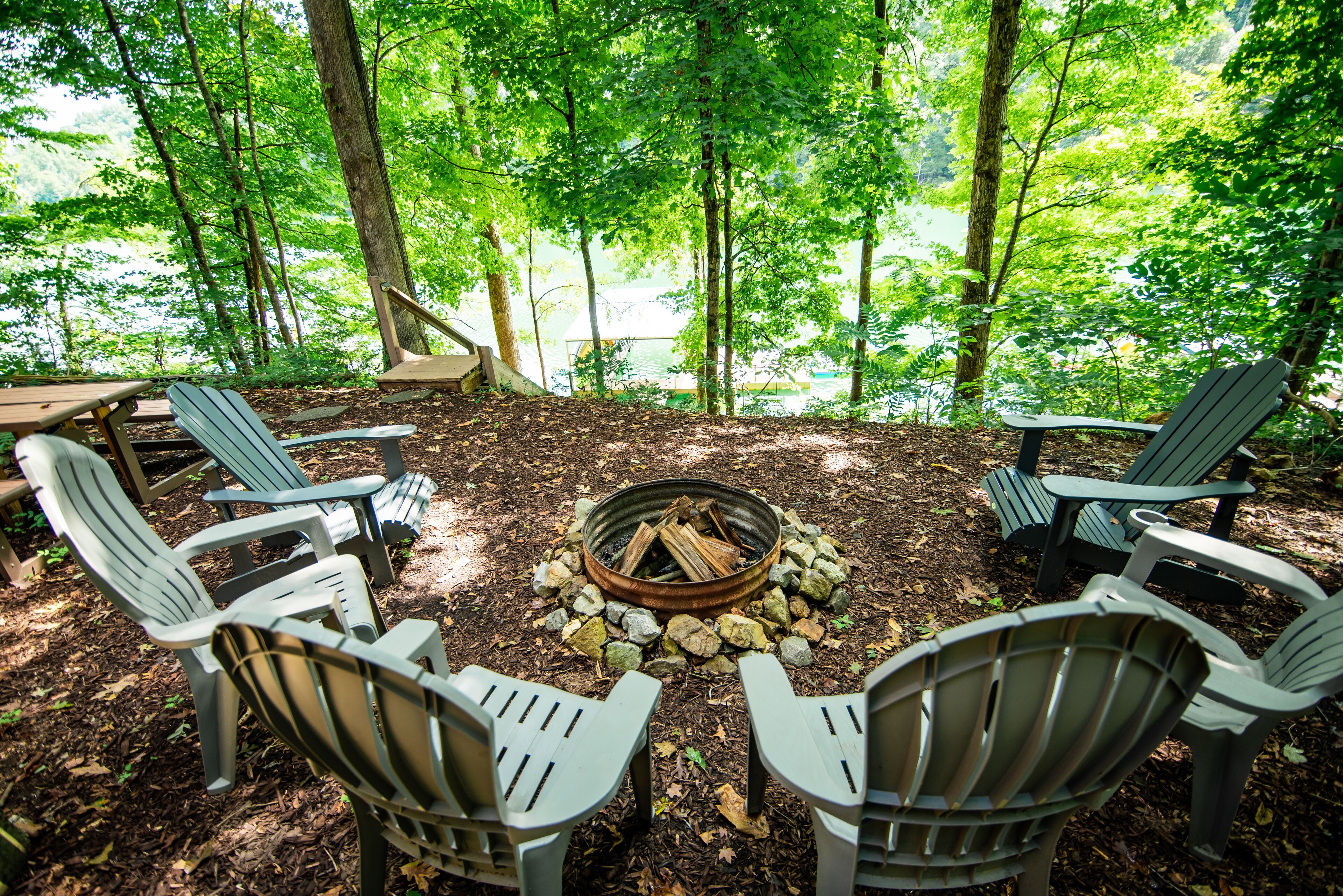 A circular fire pit surrounded by Adirondack chairs in a forested area with a view of a lake.