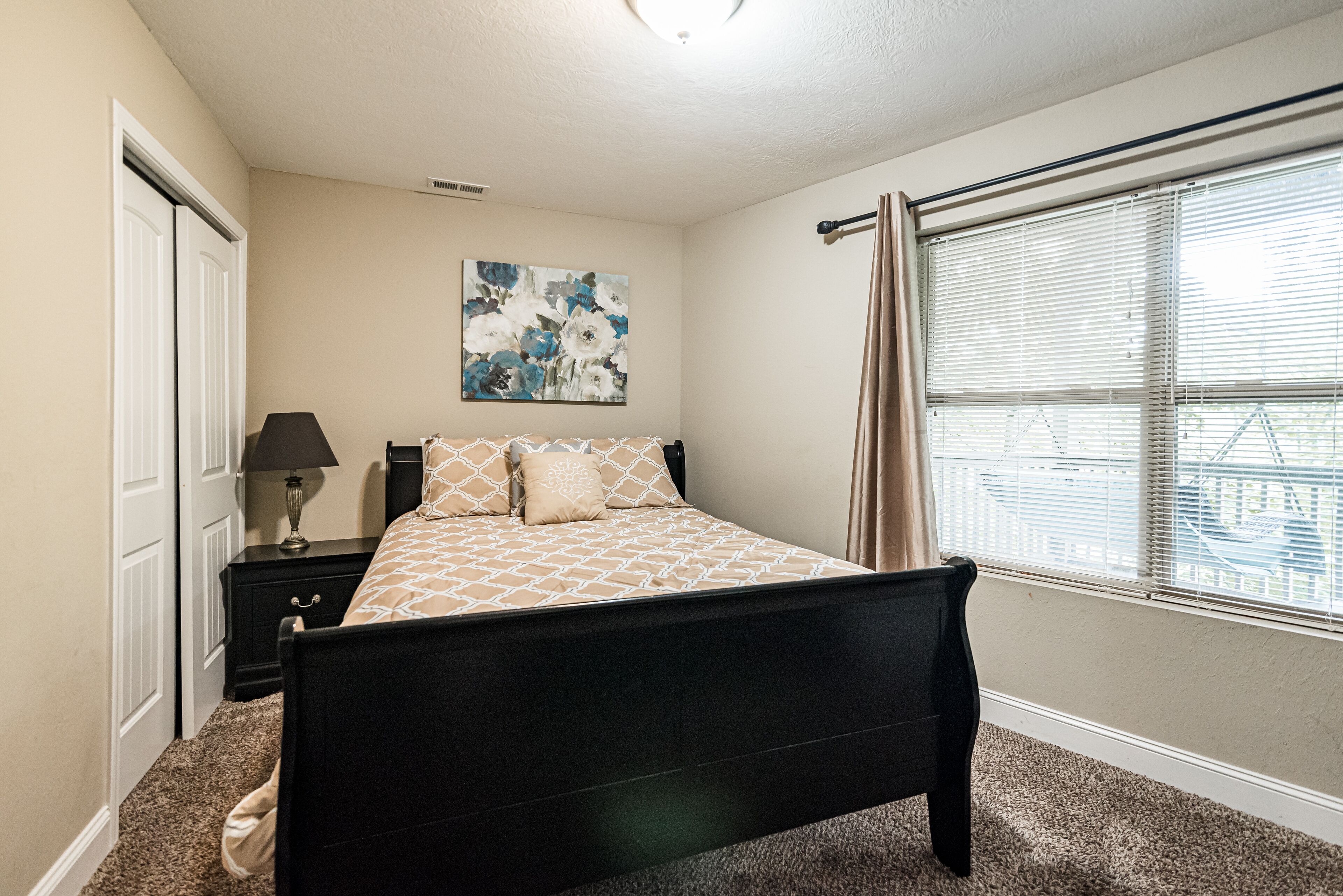 A bedroom with a bed, a nightstand, a lamp, a window with blinds, and a door.