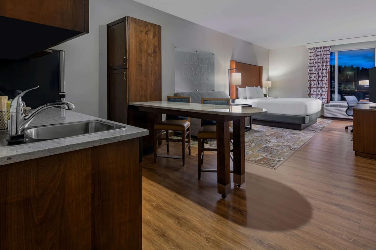 A modern hotel room with a kitchen area, a dining table, a bed, and a large window.