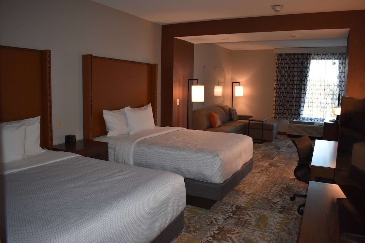 A hotel room with two beds, a desk, a chair, a sofa, and a window with patterned curtains.