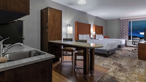 A hotel room with a kitchenette, a bed with a headboard, a desk with a chair, and a large window with a view of the city at night. - La Quinta Inn & Suites by Wyndham Flagstaff East I-40 (Flagstaff)