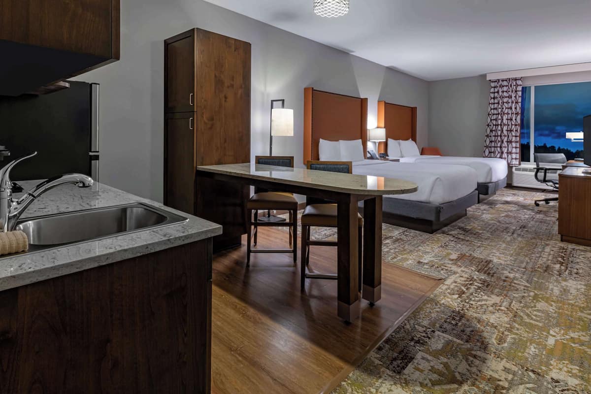 A hotel room with a kitchenette, a bed with a headboard, a desk with a chair, and a large window with a view of the city at night.