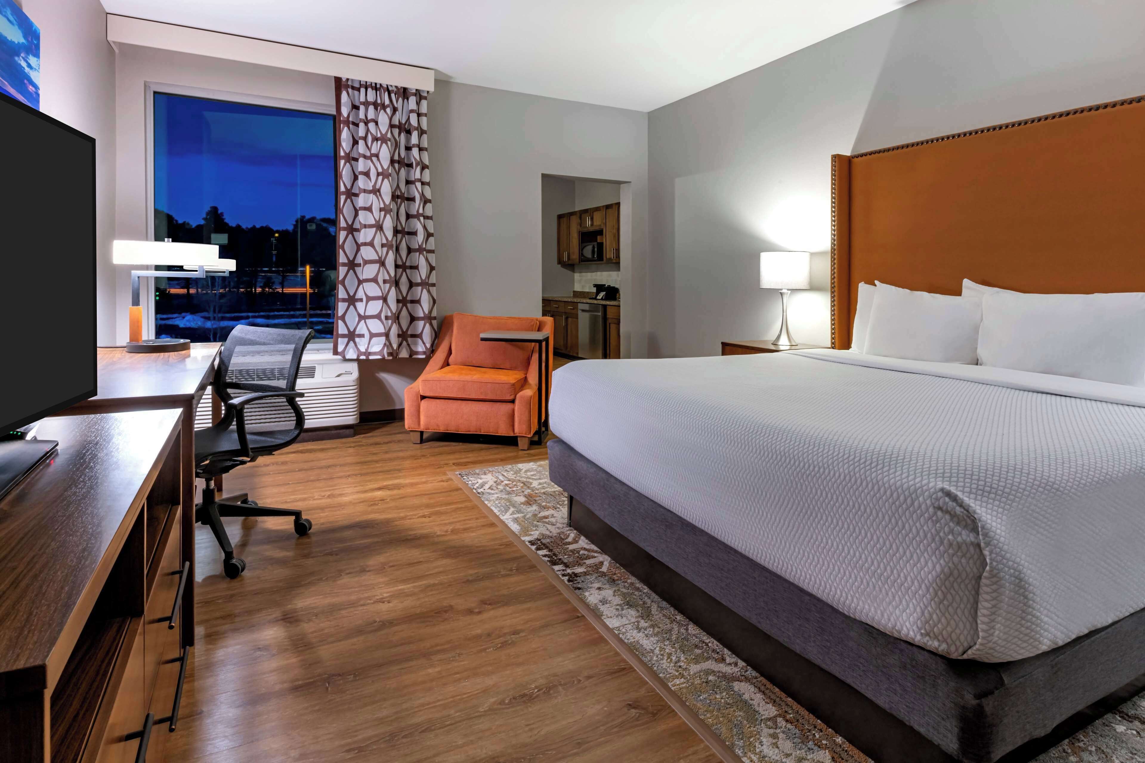 A hotel room with a large bed, a desk with a computer, an orange armchair, a view of the cityscape at dusk, and a kitchenette in the background.