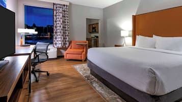 A hotel room with a large bed, a desk with a computer, an orange armchair, a view of the cityscape at dusk, and a kitchenette in the background.
