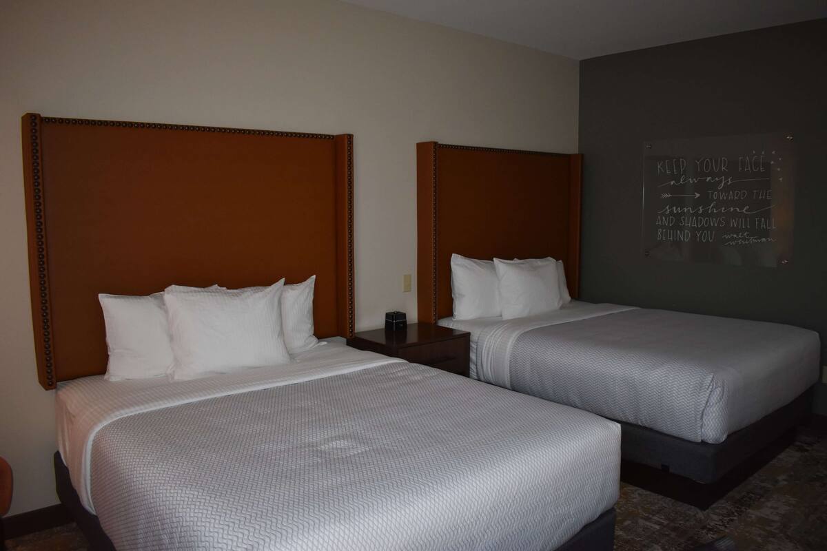 A hotel room with two beds, each with a wooden headboard, white bedding, and a bedside table with a lamp. A motivational quote is displayed on the wall.