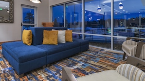 A modern hotel lobby with a blue sofa, patterned carpet, and large windows offering a view of the outdoors. - La Quinta Inn & Suites by Wyndham Flagstaff East I-40 (Flagstaff)
