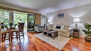 Un vaste espace de vie doté d’un plancher en bois, d’un canapé, d’une table basse et d’une table à manger avec des chaises. Deux lampes sont posées sur des tables d’appoint, et les fenêtres offrent une vue sur la verdure.