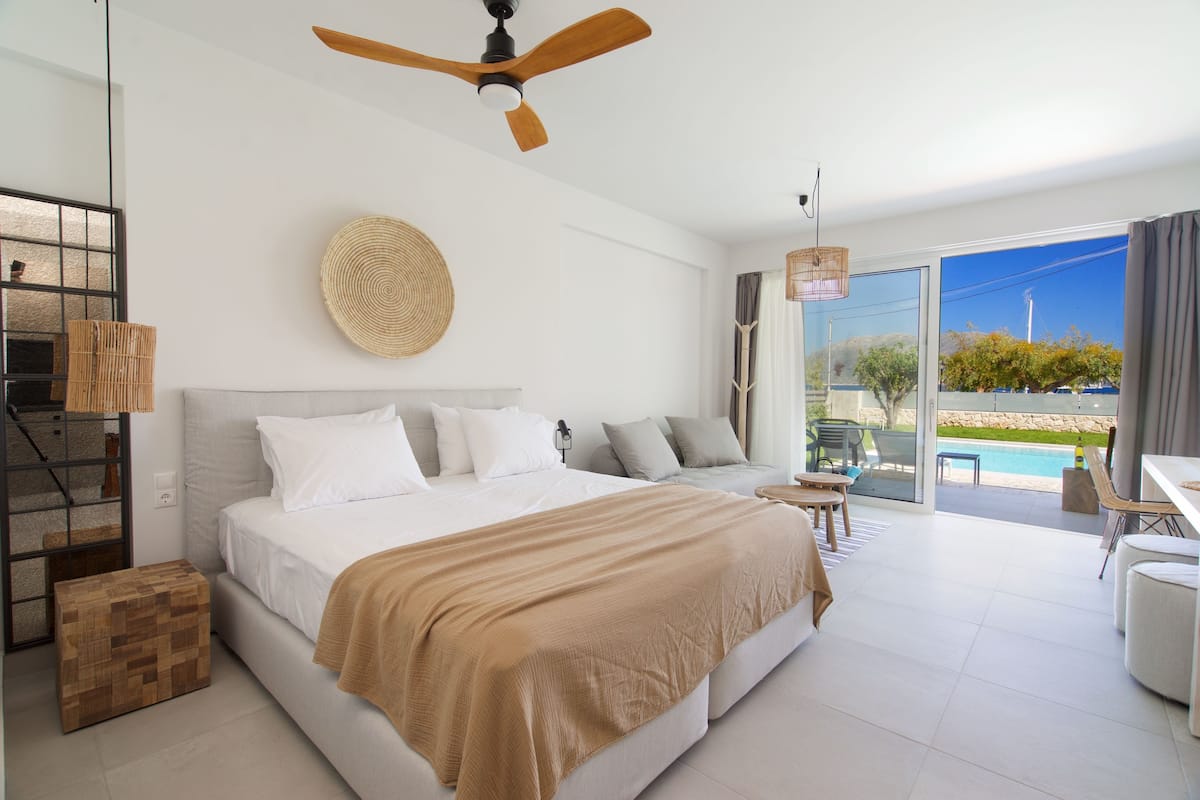 A modern bedroom with a large bed, a ceiling fan, a view of the outdoors, and a small table with chairs.