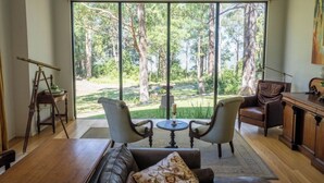 Quarto com uma grande porta de vidro deslizante que dá para uma vista da floresta. No interior, há uma mesa de madeira com duas cadeiras, um sofá de couro e uma escrivaninha de madeira com uma luminária.