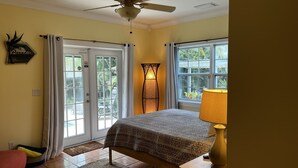 A bedroom with a bed, a ceiling fan, a nightstand with a lamp, a window with a view of greenery, and a door with a glass panel. - Melbourne Beach getaway, Surfing, Birdwatching, nature. fishing. (Melbourne Beach)