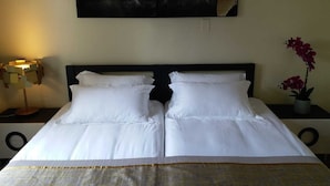 Un lit double avec du linge de lit blanc, une tête de lit sombre, un tableau représentant un léopard accroché au mur, une lampe sur pied et un orchidée en pot.