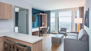 Ein modernes Hotelzimmer mit Schreibtisch, grauem Sofa, großem Fenster mit Stadtblick und Kleiderschrank.