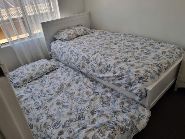 Un lit simple avec une literie à motifs floraux, dans une chambre dotée d’une fenêtre et d’une tête de lit blanche.