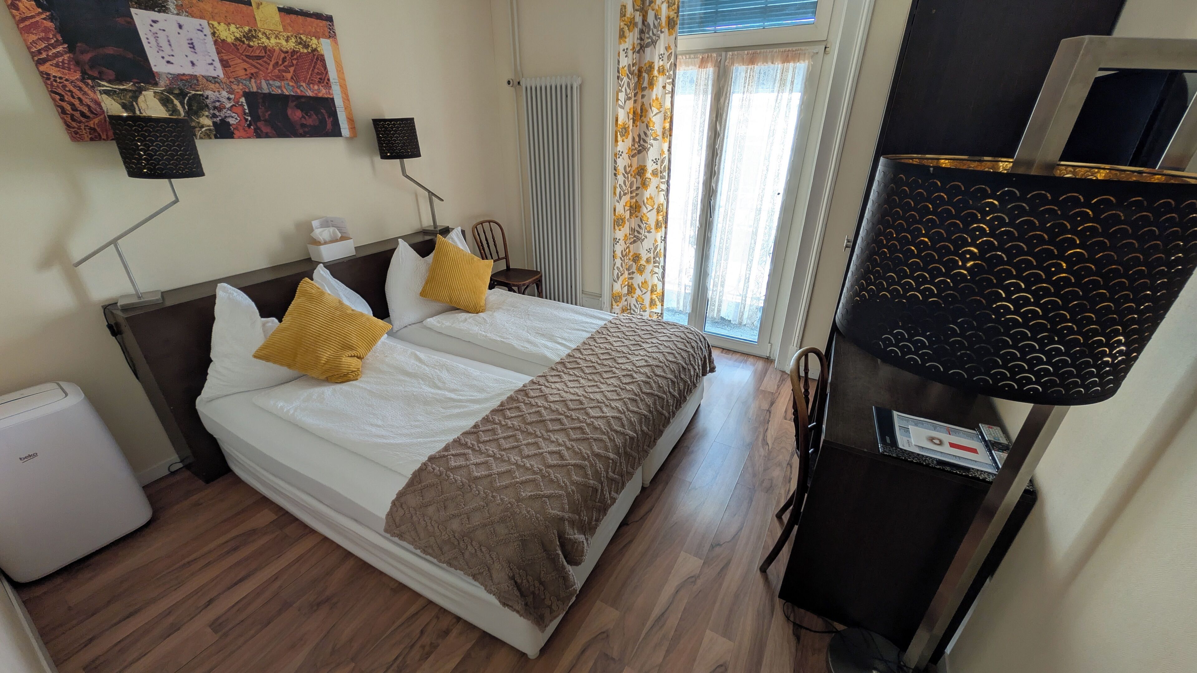Habitación de hotel con cama, escritorio, silla, radiador, ventana con cortinas y decoración en la pared.