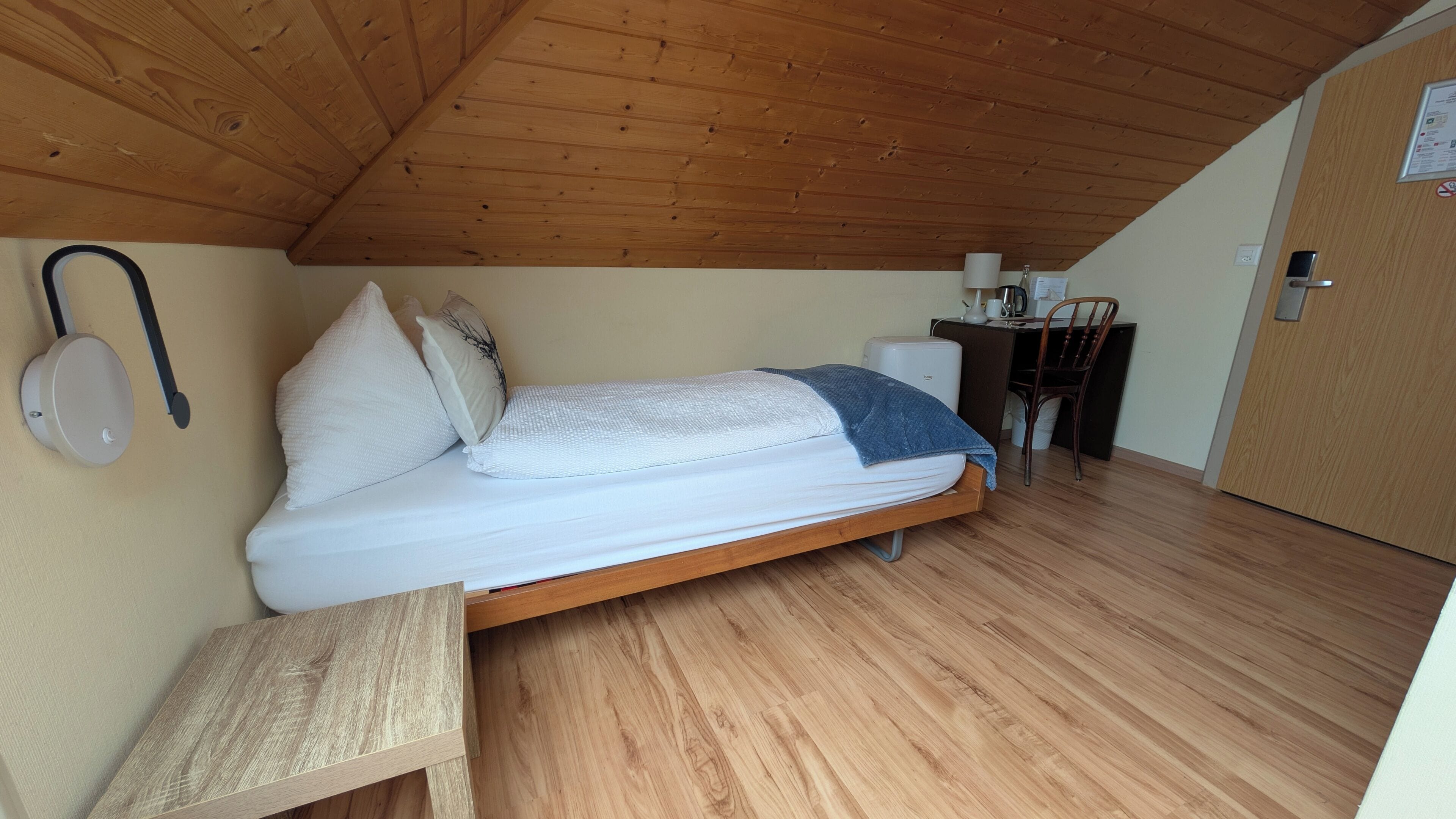 Une petite chambre avec un lit simple, un plafond et des murs en bois, un bureau avec une chaise et une petite table.