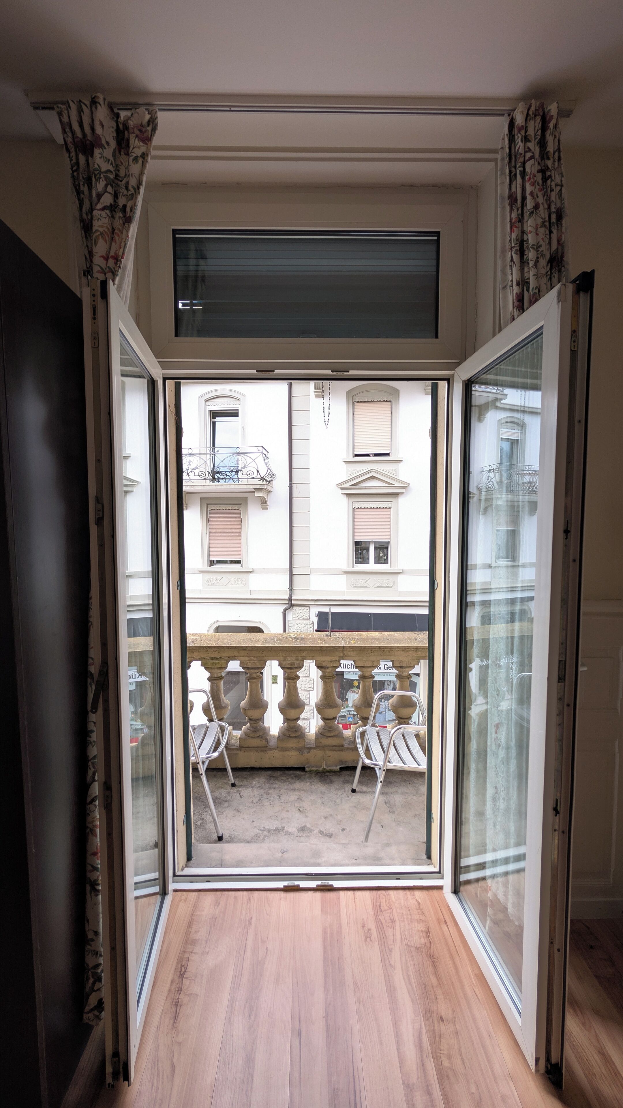 A room with a wooden floor, a sliding glass door leading to an outdoor area with a stone balustrade and two white chairs.