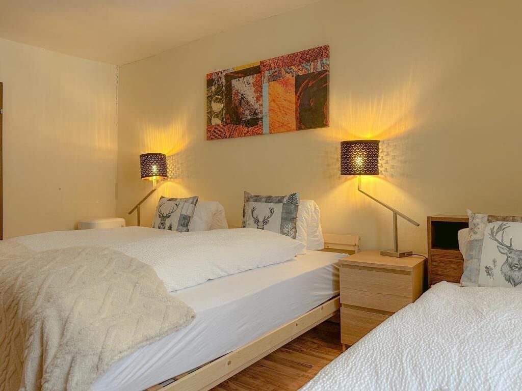 Une chambre avec deux lits, chacun doté d’un cadre en bois et de draps blancs. On y trouve deux tables de chevet avec des lampes et des coussins décoratifs. Un tableau abstrait aux couleurs vives est accroché au mur, au-dessus des lits.
